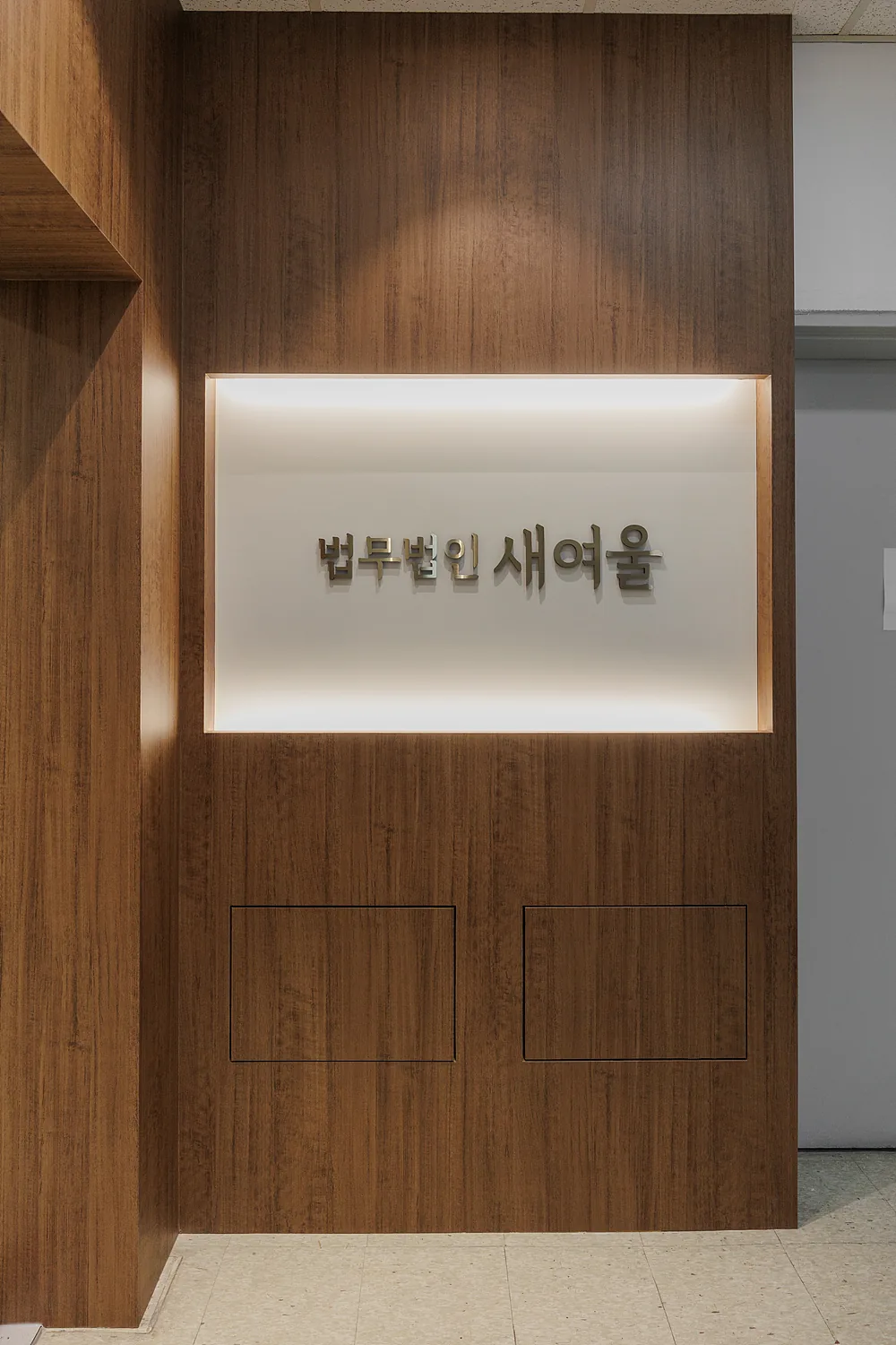 나무 벽면의 조명 박스 안에 법무법인 새여울 이름이 금속 글자로 붙어 있는 사무실 입구